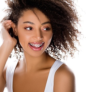 A young female wearing a white tank top and pushing her hair back while smiling and showing off her traditional braces